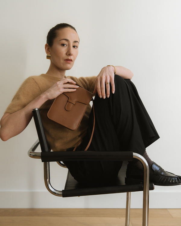 Woman sitting on a chair holding a brown leather bag against a plain background