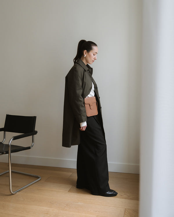 Person wearing a long dark coat and black pants, wearing a bag, standing against a plain wall with a chair in the corner.