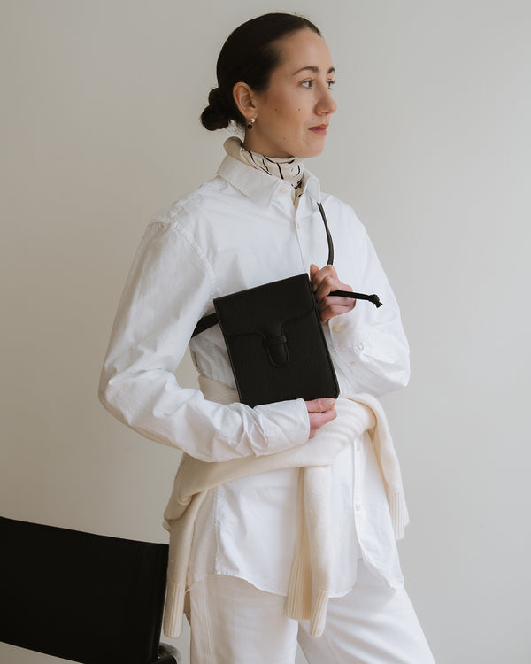 Woman in a white outfit holding a black leather phone bag against a plain background