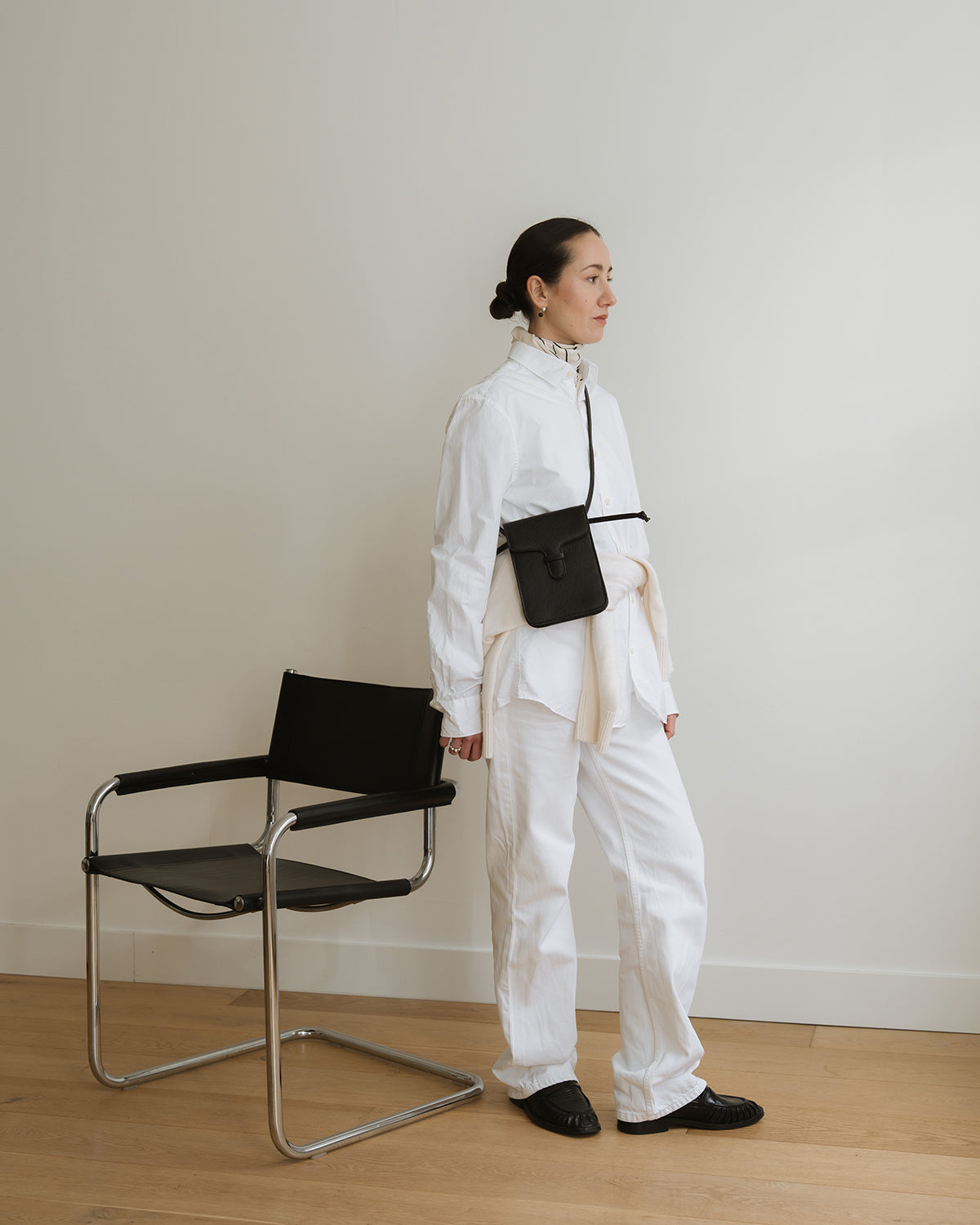 Person wearing a white outfit with a black bag standing next to a modern black chair against a plain wall.