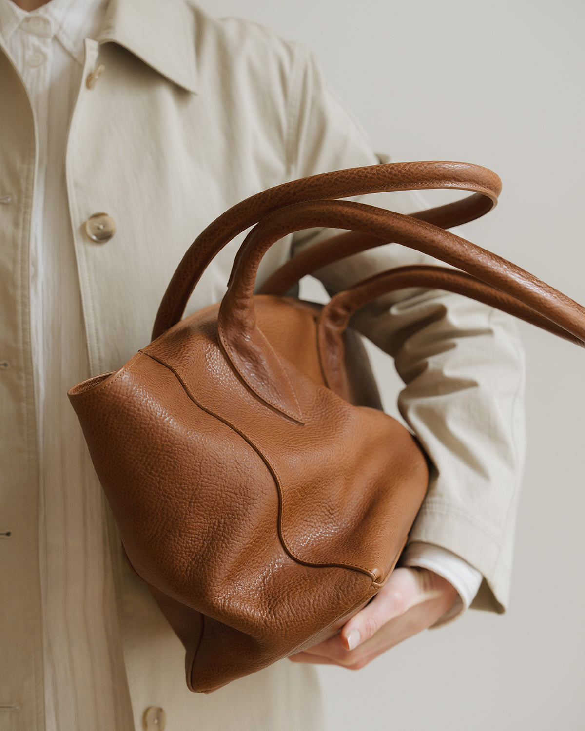Brown leather handbag held by a person wearing a beige coat.
