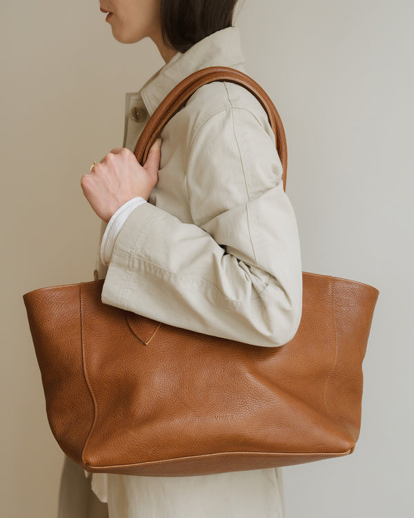 Person holding a brown leather tote bag against a neutral background